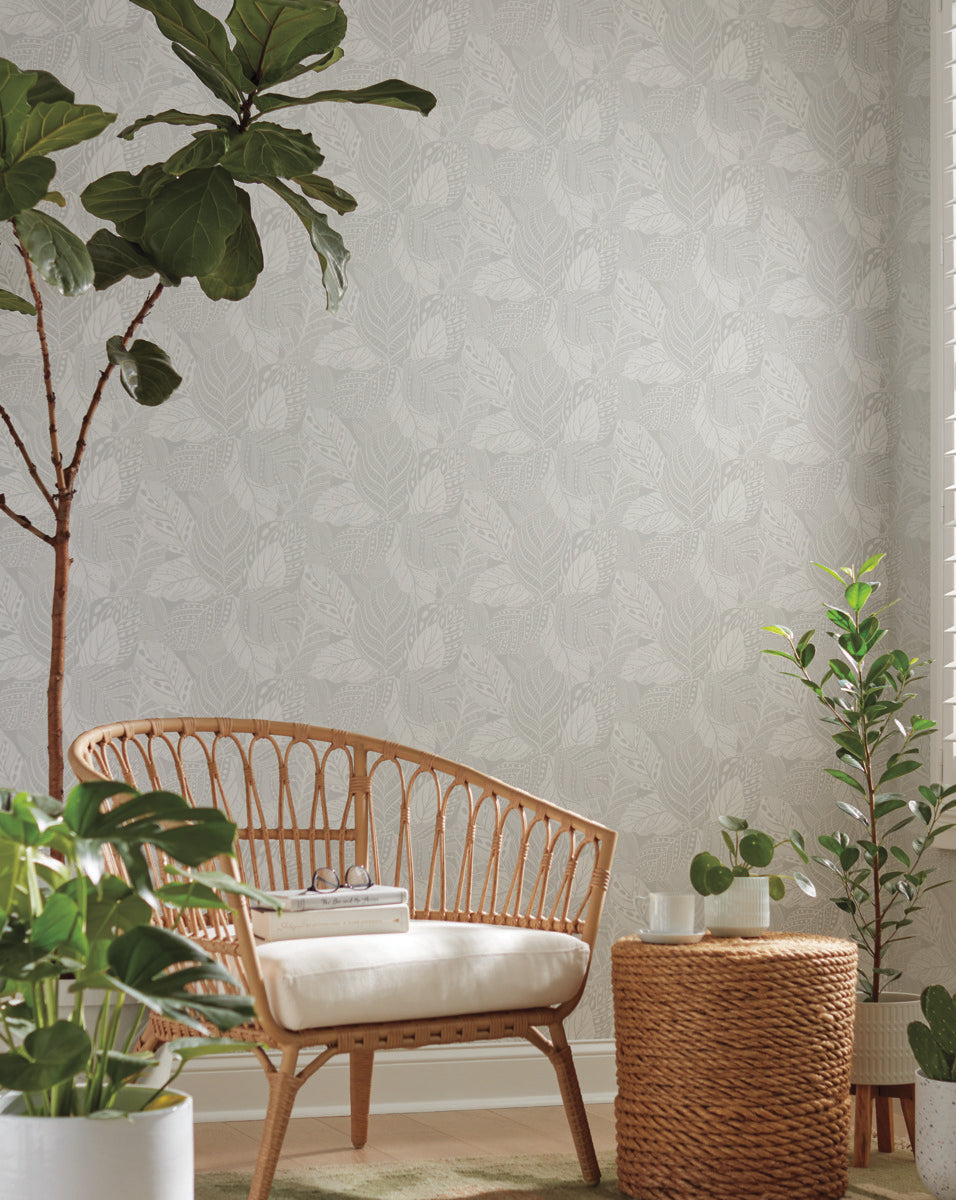 A cozy room decorated with multiple green plants, featuring a wicker chair with a white cushion, a round wicker side table, and York Wallcoverings Vinca Glint Wallpaper Metallic (60 Sq.Ft.) with a subtle leaf pattern. Sunlight streams in from the right, brightening the space.