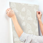 A person with a striped long-sleeve shirt is applying a York Wallcoverings Vinca Glint Wallpaper Metallic (60 Sq.Ft.) to a white wall. The wallpaper, made from durable non-woven material, features an intricate design of overlapping leaves in neutral tones. The person's hands are smoothing out the wallpaper as it is being applied.