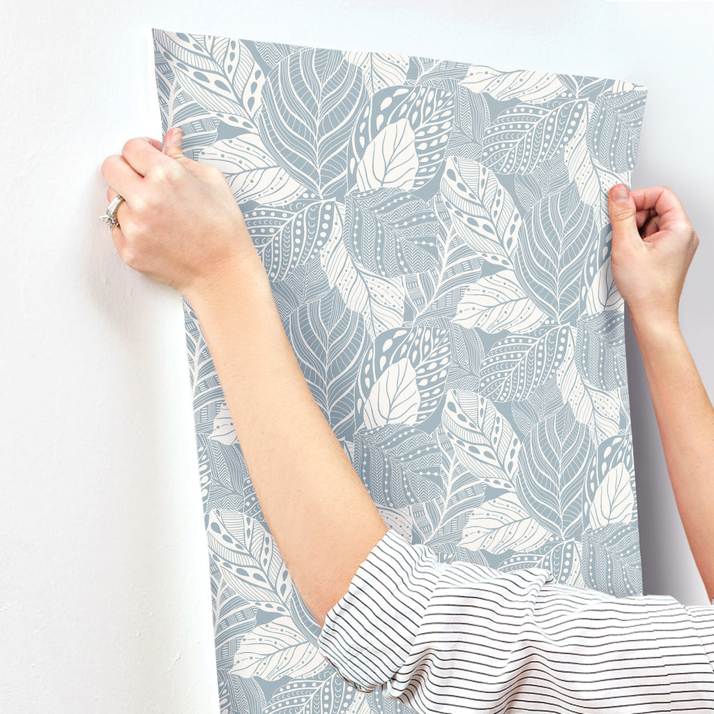 A person is applying Vinca Eucalyptus Wallpaper Green (60 Sq.Ft.) by York Wallcoverings on a wall. The wallpaper features a detailed leaf design in shades of blue and grey. Only their arms are visible.