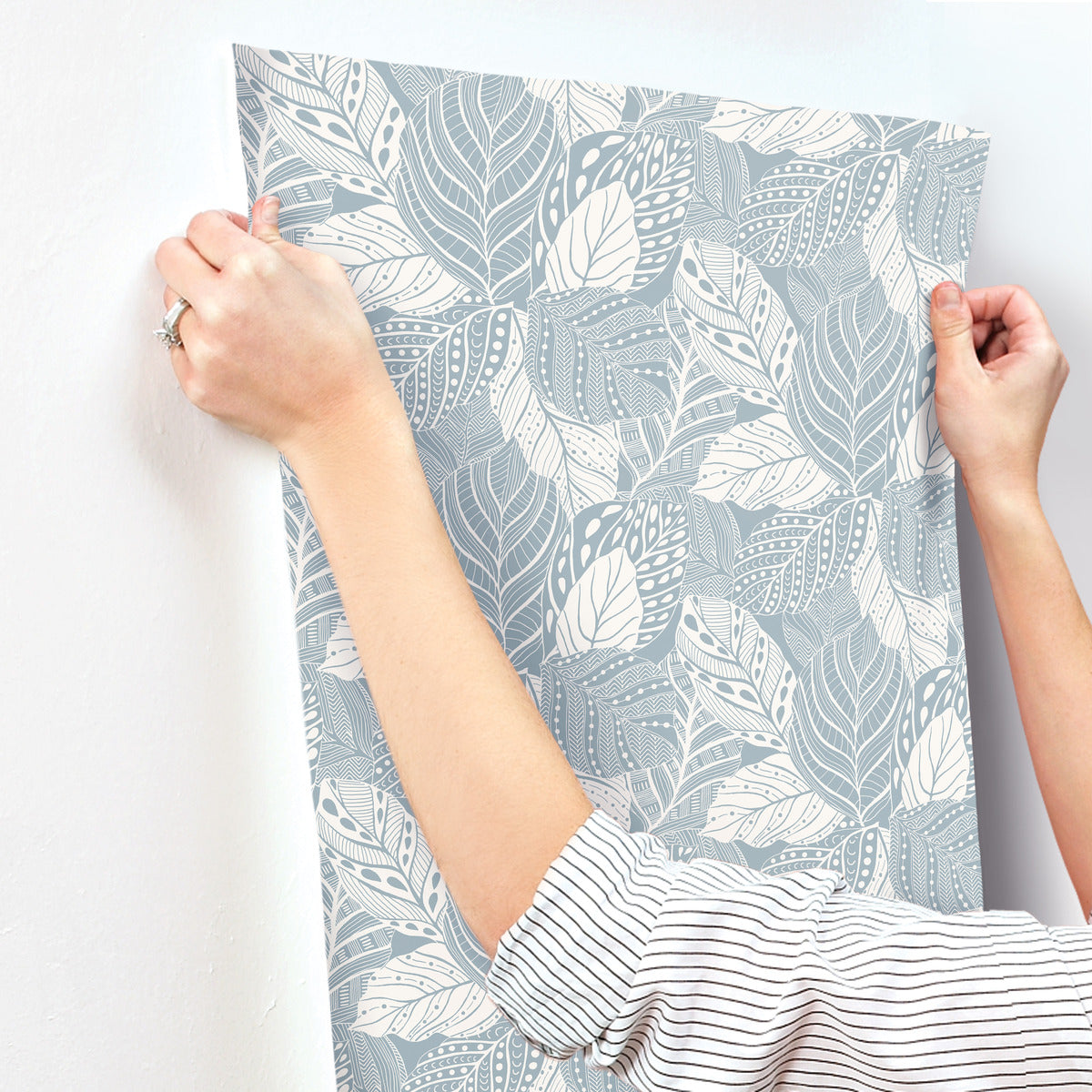 A person is aligning and attaching a piece of Vinca Smokey Blue Wallpaper Blue (60 Sq.Ft.) by York Wallcoverings, featuring a pattern of white leaves on a light blue background, to a white wall. Wearing a long-sleeved striped shirt and a ring on their finger, they appreciate the durable material that makes it easy to hang and remove.