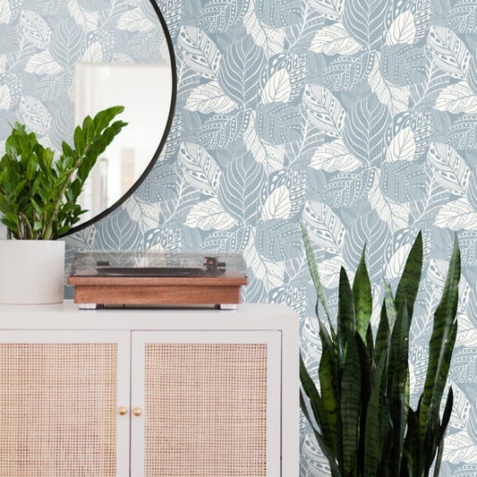 A minimalist interior features a white cabinet adorned with a potted plant and a record player. A round mirror reflects part of the room, and a snake plant stands next to the cabinet. The wall is decorated with Vinca Smokey Blue Wallpaper Blue (60 Sq.Ft.) by York Wallcoverings in blue and white leaf patterns, making redecorating effortless.