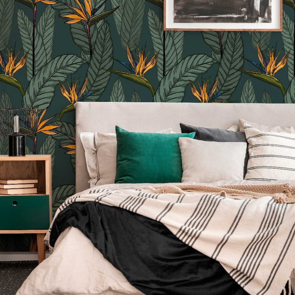 A cozy bedroom corner with a bed covered in a striped blanket and various pillows, beside a wooden nightstand under a lamp, against Decor2Go Wallpaper Mural with floral designs. A framed picture hangs above.