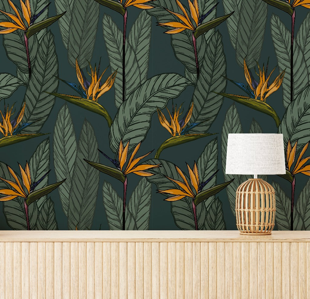 A Decor2Go Wallpaper Mural Vintage Botanical Garden Wallpaper Mural features large green leaves and vivid orange bird of paradise flowers. In front of the wallpaper is a wicker table lamp with a white shade, placed on a wooden table with vertical paneling, adding a touch of nostalgia to the interior design.
