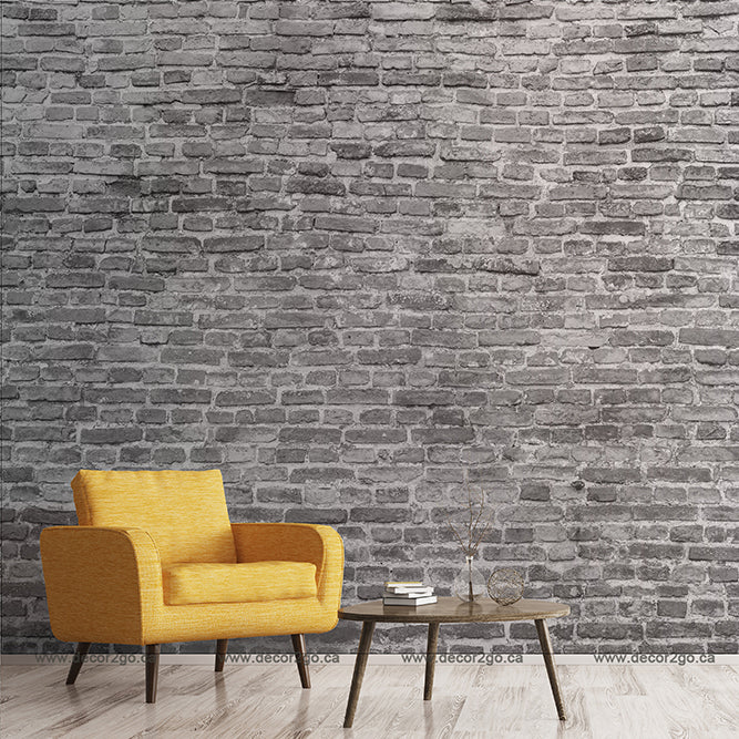 A contemporary room featuring a Decor2Go Washed Grey Brick Wall Wallpaper Mural with realistic details, complemented by a vibrant yellow armchair to the left, a small round wooden table with books and a plant in the center.