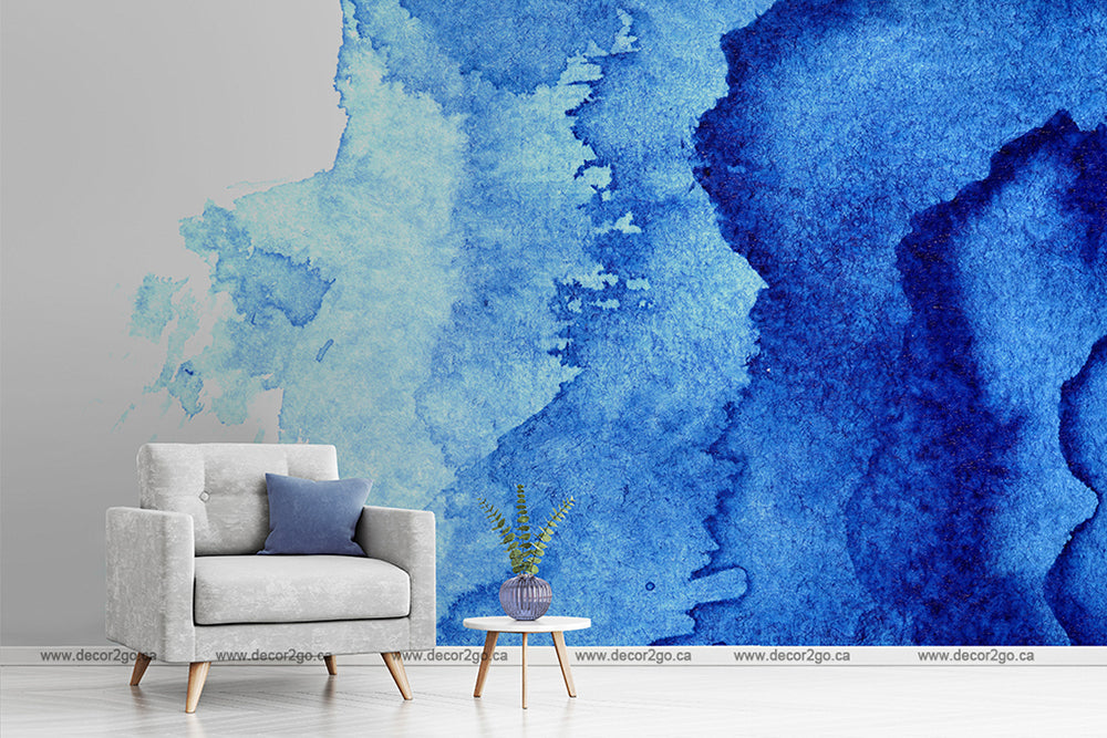 A modern artist's space featuring a light grey armchair with blue pillows beside a small deep blue side table with a plant, set against a large Decor2Go Wallpaper Mural with abstract Waterblue Ink strokes.