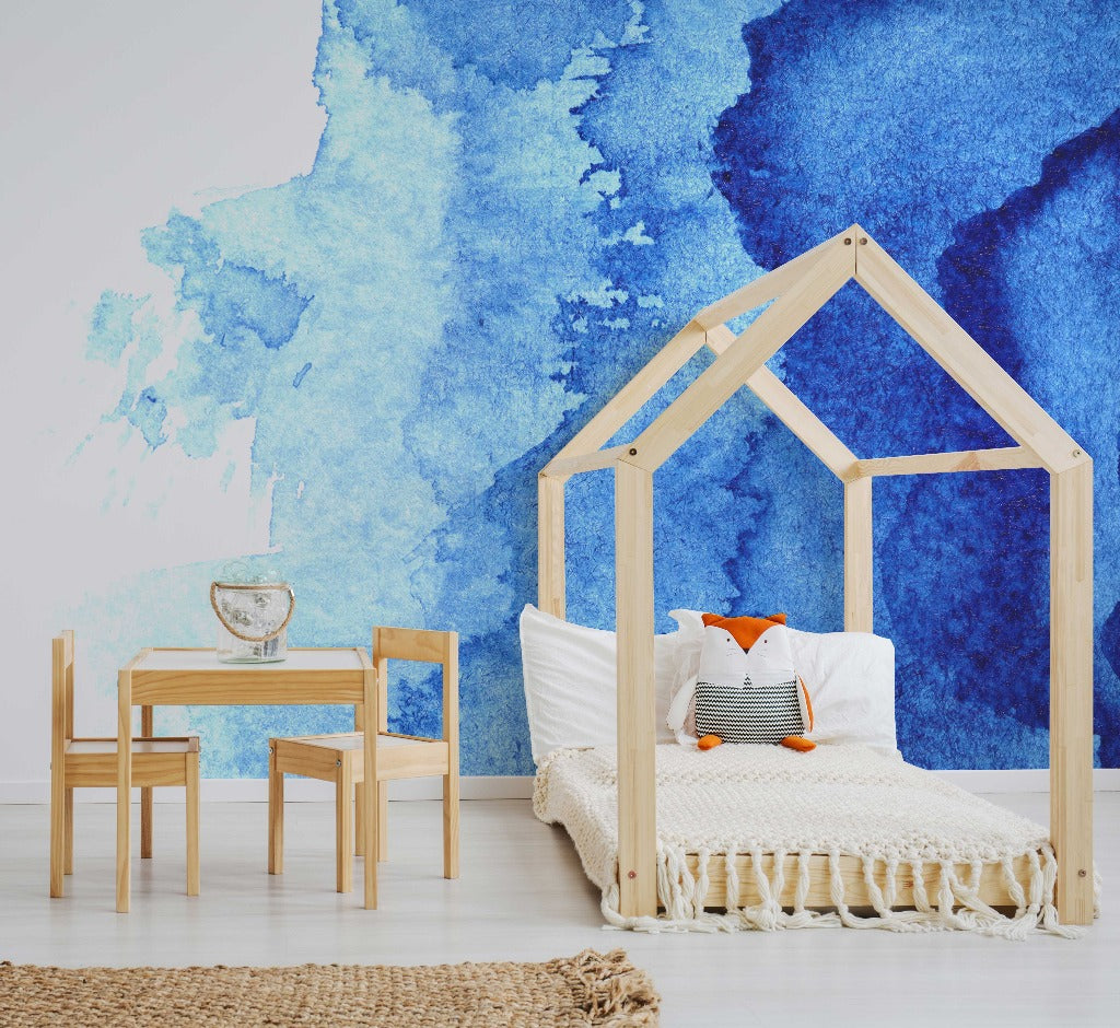 A stylish children's bedroom featuring a wooden house-shaped bed frame with a white mattress, orange pillow, and a fish-shaped pillow. There's a small wooden table and chairs on a rug, next to Decor2Go Wallpaper Mural's Waterblue Ink Wallpaper Mural.