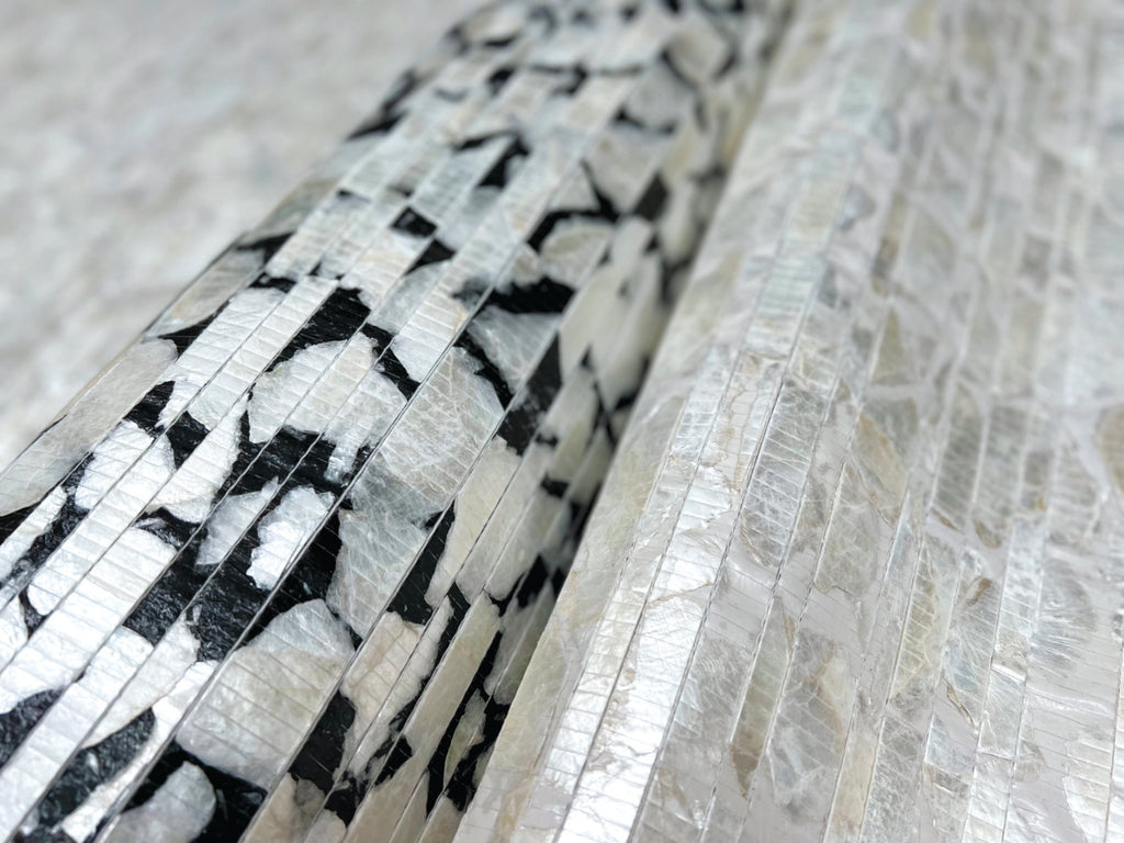 Close-up view of a cylindrical object with a patterned black and white York Wallcoverings White Pearl Capiz Offering Wallpaper (60 sq.ft.) surface, lying on a textured silver-white metallic background.