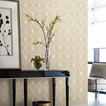 In a living room corner, a black console table with a glass vase featuring a flowering branch stands beside a decorative ball dish. A framed abstract art piece hangs above, enhanced by York Wallcoverings' White & Gold Harlowe Wallpaper, while a cozy beige armchair invites relaxation.