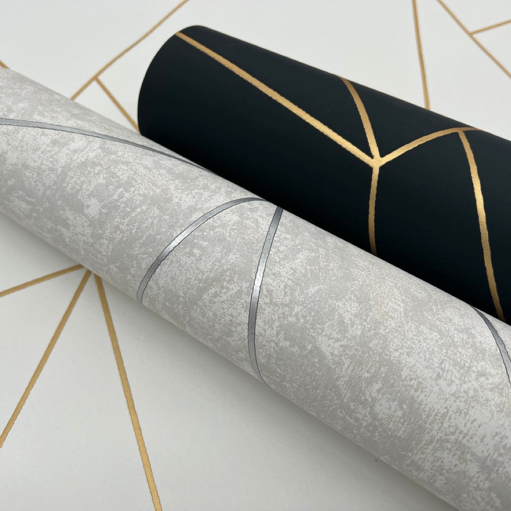 Two rolls of York Wallcoverings’ White & Gold Nazca Wallpaper (57 SqFt) are laid side by side. One is light grey with silver geometric lines and the other dark with gold geometric lines, featuring a geometric pattern from the Modern Metals Collection in the background.