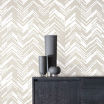 A dark wooden sideboard with three minimalist gray cylindrical vases rests against a wall covered in York Wallcoverings' White & Gold Polished Chevron Wallpaper, creating a sleek and contemporary aesthetic.