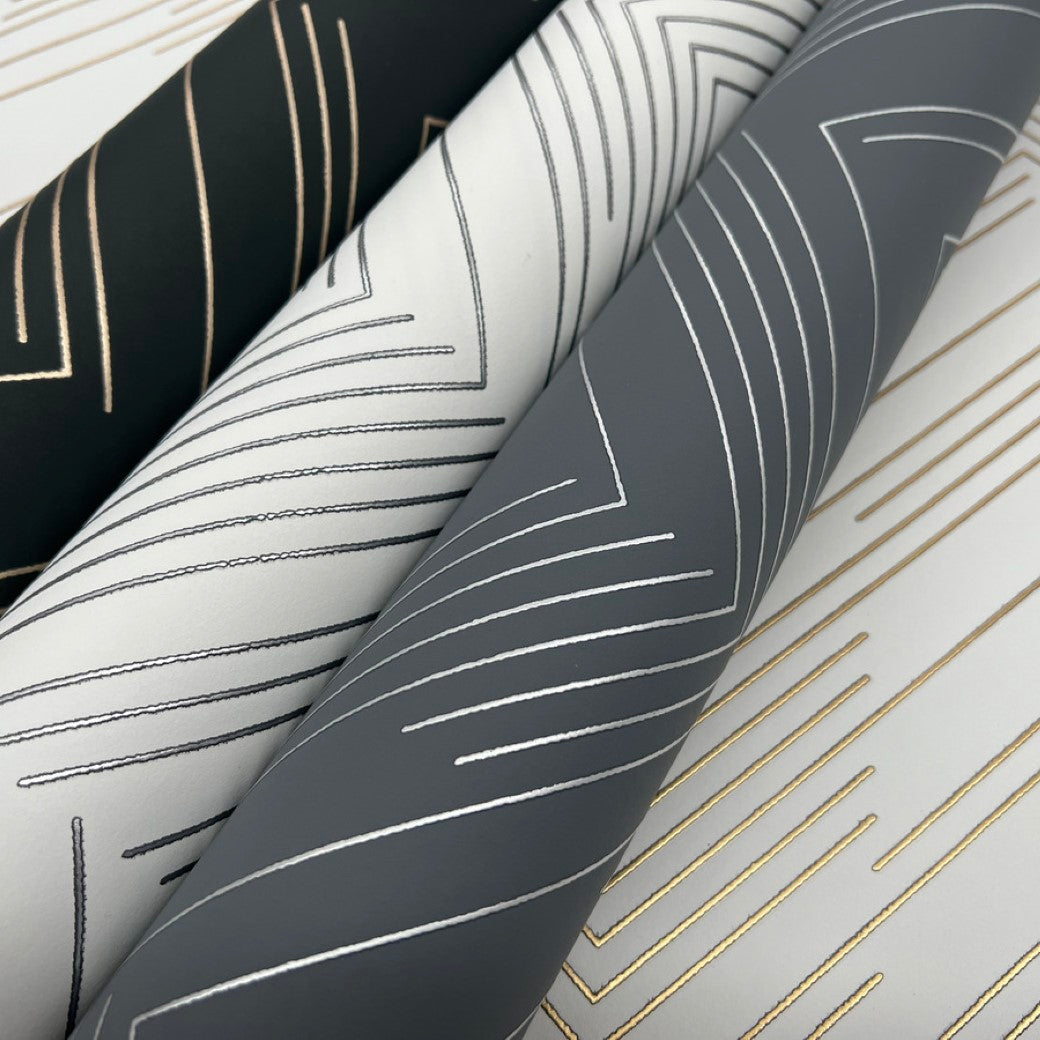 Close-up of three fabric rolls featuring geometric metallic designs with lines and angles that echo the York Wallcoverings' White & Gold Polished Chevron Wallpaper, set against black, gray, and white backgrounds.