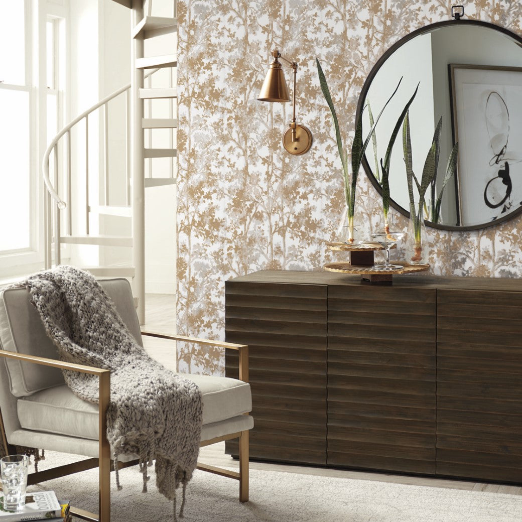 Living Room - A cozy corner features a modern armchair with a gray knit throw. A dark wood sideboard is topped with a large mirror, plants, and decor. Elegance is enhanced by York Wallcoverings' White & Gold Shimmering Foliage Wallpaper and a spiral staircase