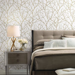 A modern bedroom features a beige upholstered bed, white and beige bedding, assorted pillows, and a sleek wooden nightstand with a lamp, books, and flowers. The York Wallcoverings White & Gold Trees Silhouette Wallpaper adds depth with its subtle branch pattern for a serene look.