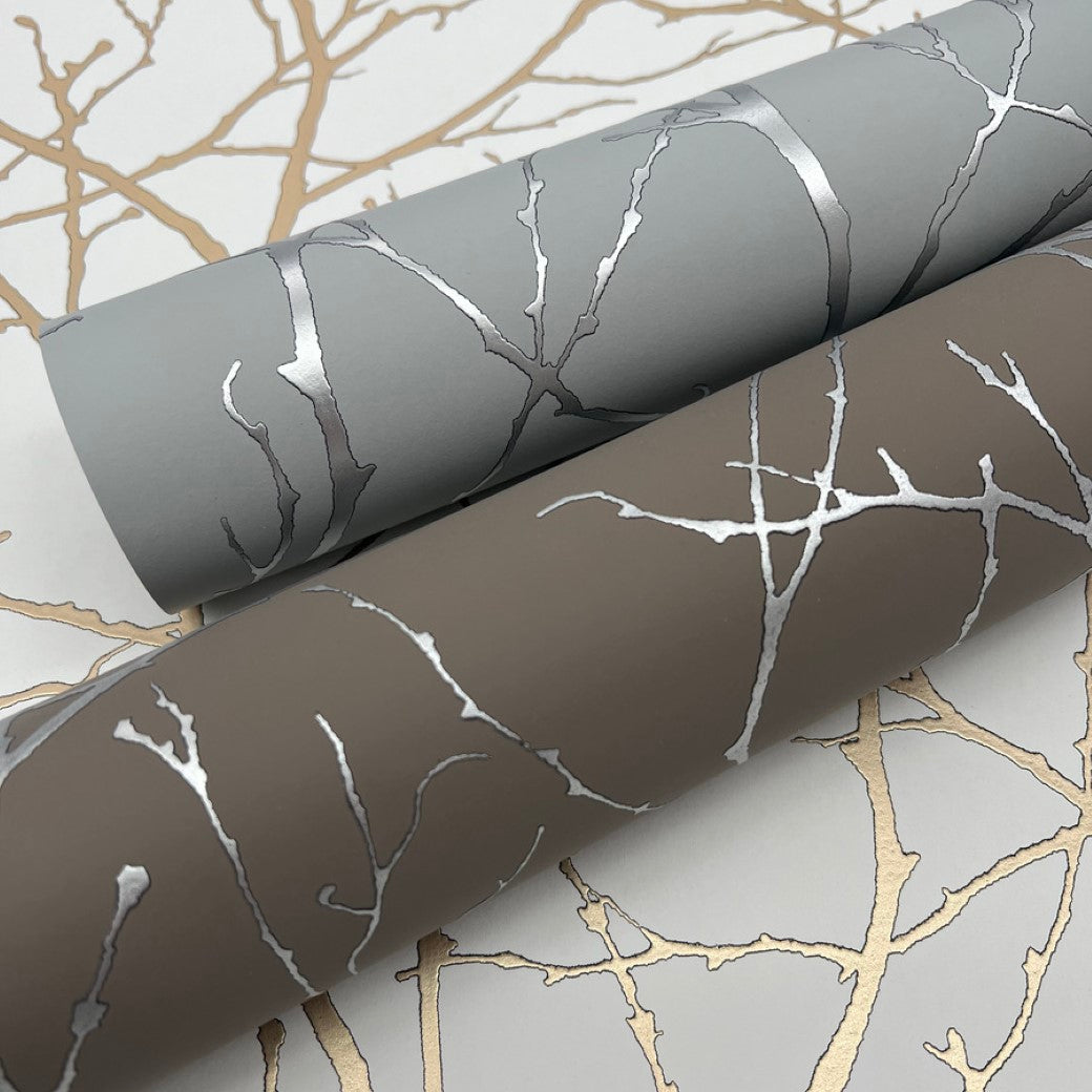 Part of York Wallcoverings' Modern Metals Collection, these two wallpaper rolls boast a metallic tree branch pattern. The first is gray with silver branches, the second brown with silver—both on light backgrounds adorned with shimmering gold branches.