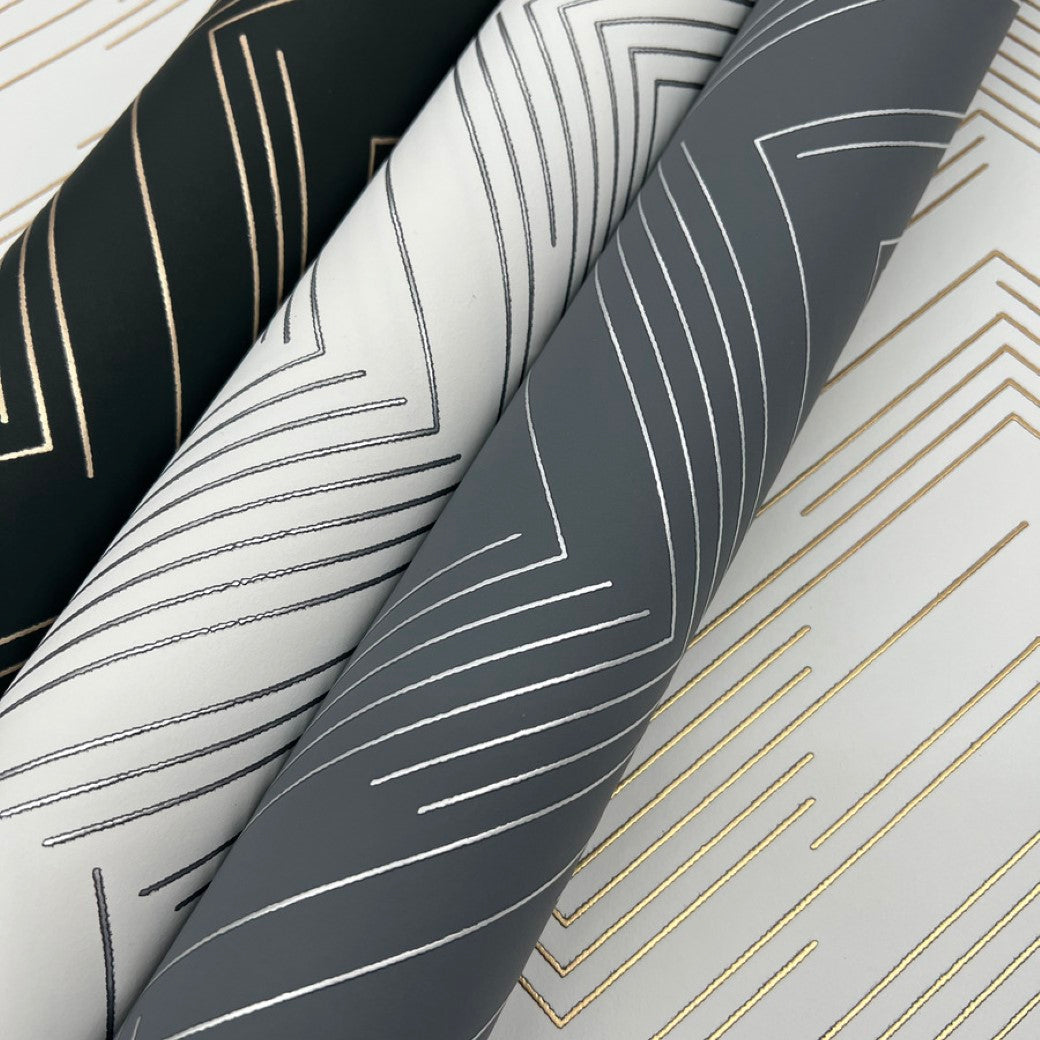 Three fabric rolls, featuring York Wallcoverings' sleek White & Silver Polished Chevron Wallpaper, display a modern metallic style in black, white, and gray with geometric accents and contrasting gold and silver lines.