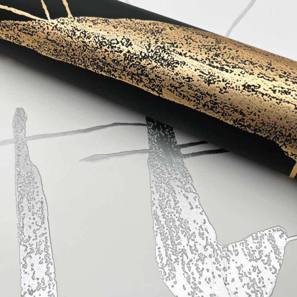 A close-up of a rolled material displays an abstract, textured gold and black design atop the White & Silver Abstract Geo Wallpaper by York Wallcoverings; a gray and white patterned background adds a modern touch, ideal for contemporary interiors.