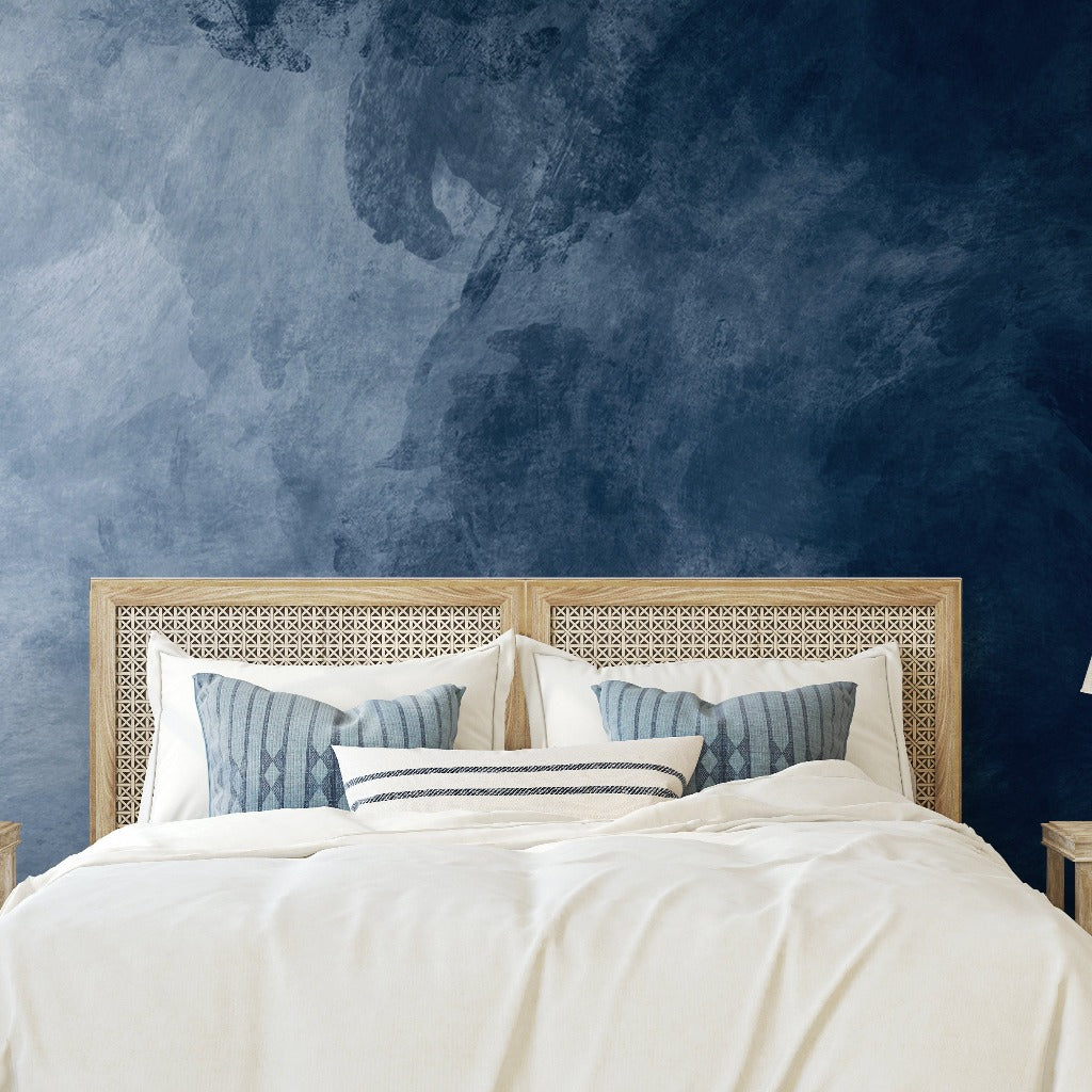 A modern bedroom featuring a bed with a decorative beige headboard and white bedding, complemented by blue and white pillows, against an Decor2Go Wallpaper Mural deep blue textured feature wall.