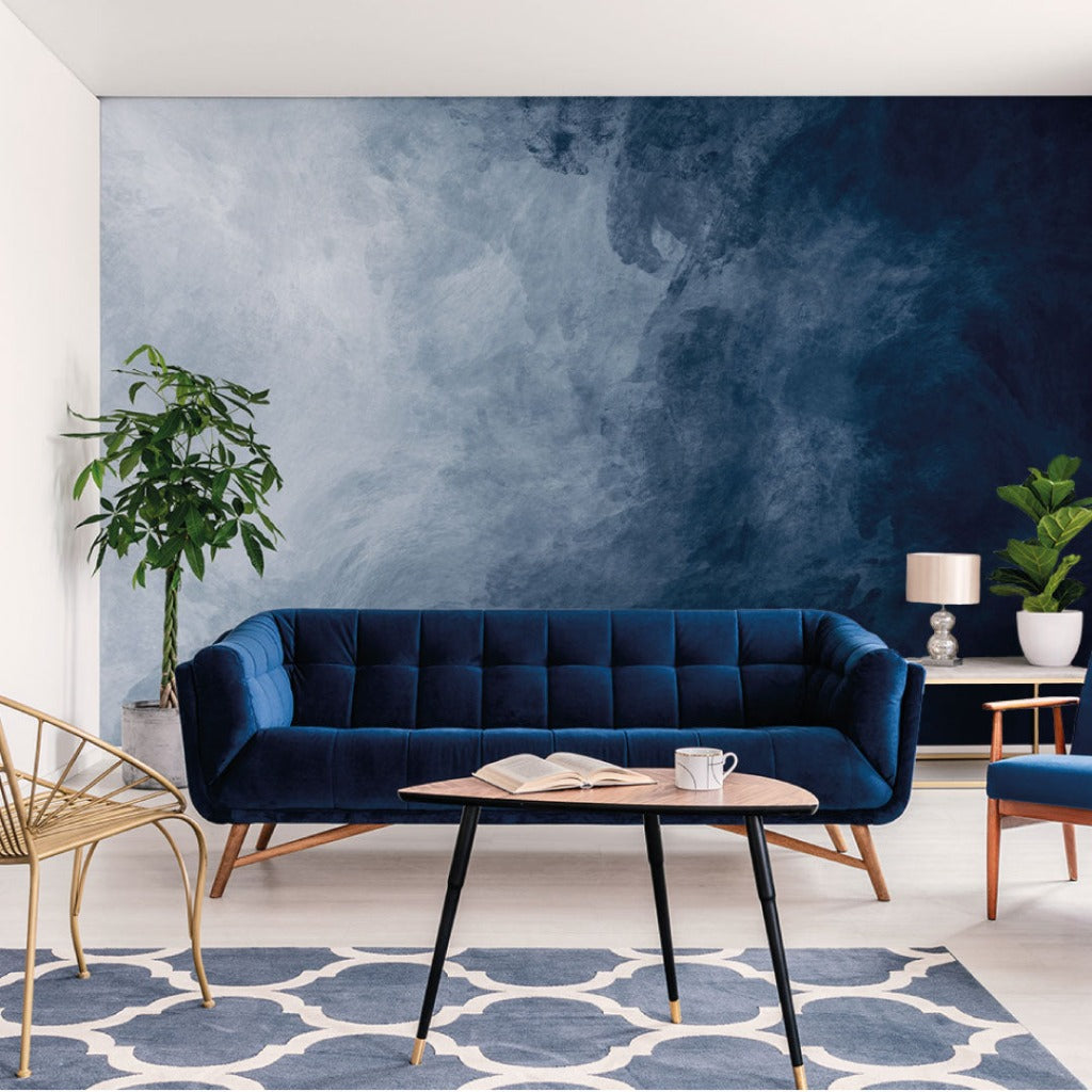 A modern living room featuring a dark blue velvet sofa against an Abstract Decor2Go Wallpaper Mural, a wooden coffee table with books and a cup, a golden chair, and a potted plant.