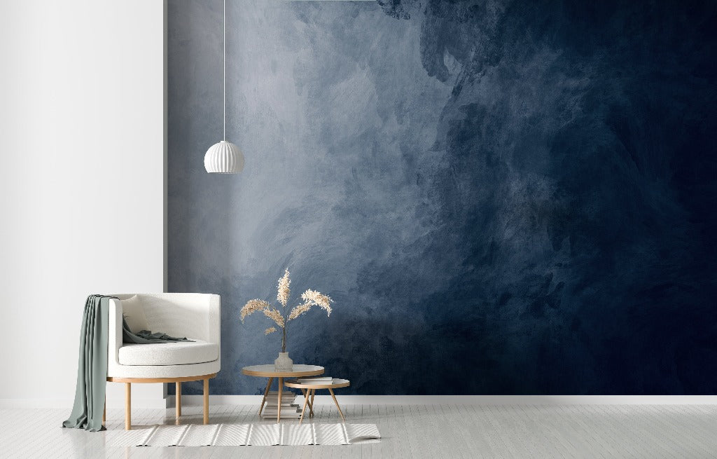 A modern living room corner with a white armchair, a wooden side table with books and a vase with dried flowers, against a textured deep blue feature wall. A simple white pendant lamp hangs above the Decor2Go Wallpaper Mural.