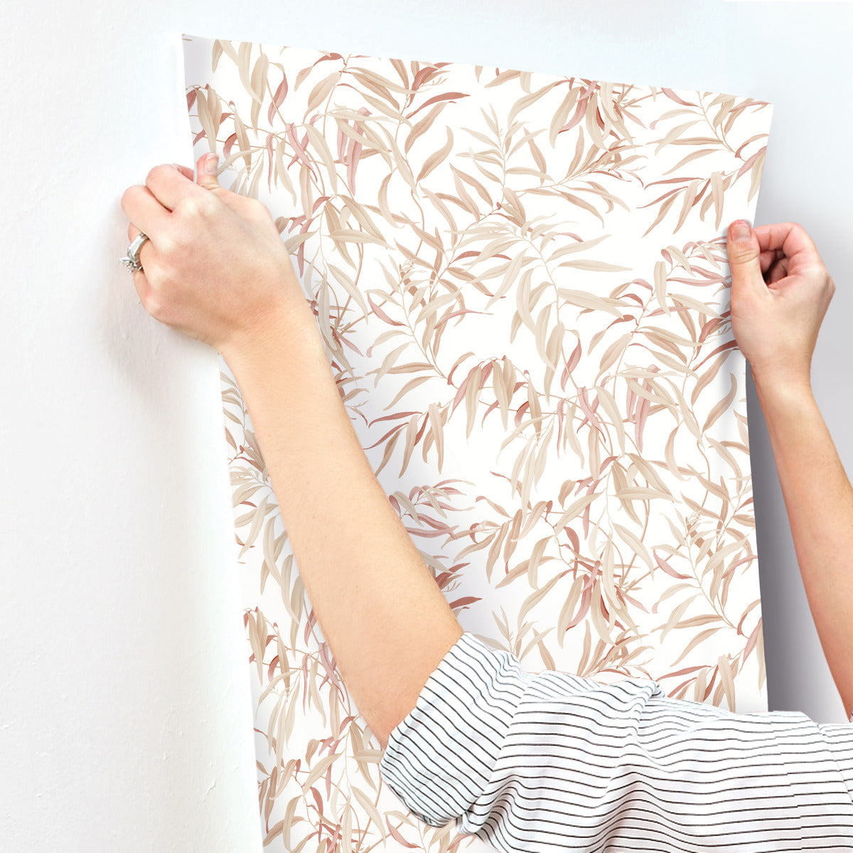 A person with a striped shirt uses both hands to adhere a sheet of York Wallcoverings Willow Grove Sand Wallpaper Beige, Grey (60 Sq.Ft.) with a delicate leaf pattern on a white wall. The wallpaper, showcasing beige and light pink leaves, exudes natural elegance as the person adjusts it to ensure it's smooth and properly aligned.