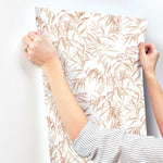 A person with a striped shirt uses both hands to adhere a sheet of York Wallcoverings Willow Grove Sand Wallpaper Beige, Grey (60 Sq.Ft.) with a delicate leaf pattern on a white wall. The wallpaper, showcasing beige and light pink leaves, exudes natural elegance as the person adjusts it to ensure it's smooth and properly aligned.