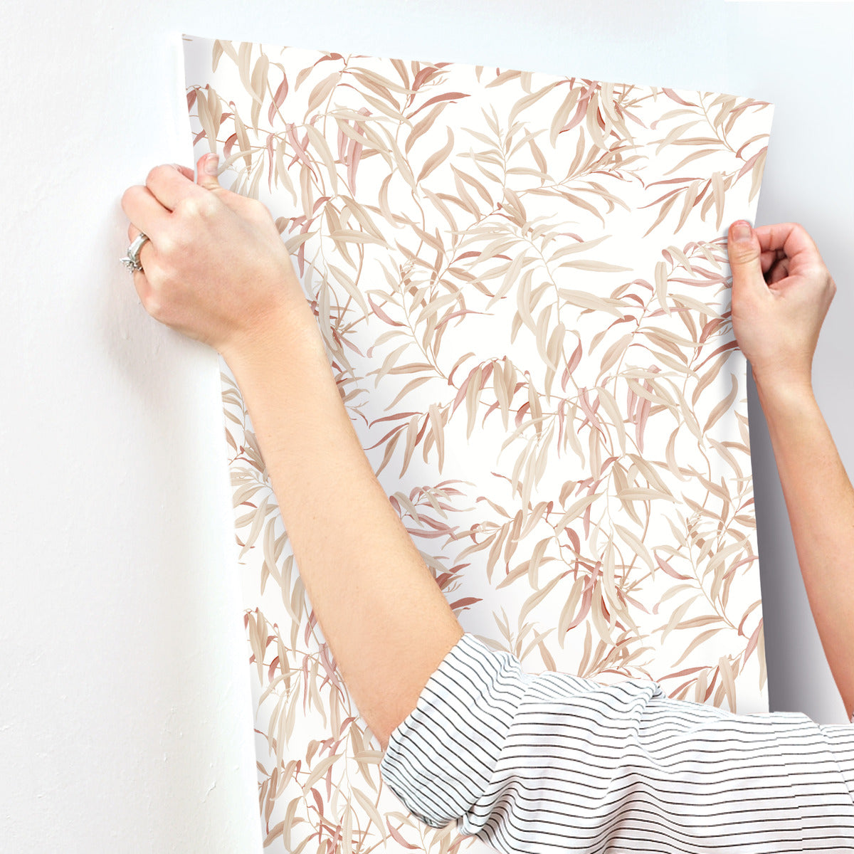A person wearing a striped shirt is applying York Wallcoverings Willow Grove Clay Wallpaper Pink (60 Sq.Ft.) with a leafy, pastel-colored botanical design to a white wall. The SureStrip wallpaper features an elegant pattern of light pink and beige leaves, creating a serene botanical retreat.