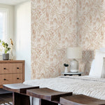 A cozy bedroom features a bed with light-colored linens, a wooden bench at the foot, a wooden dresser with a potted plant and decorative items, a bedside table with a lamp, and a window. The wall behind the bed is adorned with Willow Grove Sky Wallpaper Blue (60 Sq.Ft.) by York Wallcoverings featuring beige and brown leafy patterns.