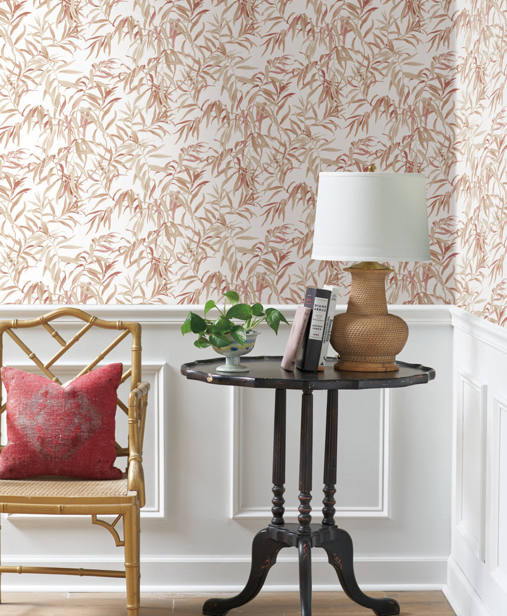 A cozy corner featuring a bamboo chair with a red cushion, a dark wooden side table holding a wicker lamp with a white shade, and a small potted plant. The backdrop of beige and grey leaf-patterned Willow Grove Sand Wallpaper Beige, Grey (60 Sq.Ft.) by York Wallcoverings, combined with white wainscoting, adds natural elegance to the space.