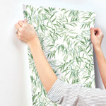 A person is hanging York Wallcoverings Willow Grove Clay Wallpaper Pink (60 Sq.Ft.) with a green leafy vine pattern on a white wall. The person has a ring on their left hand and is wearing a striped long-sleeve shirt. The SureStrip wallpaper design features intricate, delicate vines and leaves, creating a botanical retreat.