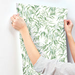 A person with a striped shirt is holding up York Wallcoverings Willow Grove Sand Wallpaper Beige, Grey (60 Sq.Ft.), featuring a green leafy pattern, against a white wall. Possibly preparing to apply the wallpaper, the person showcases natural elegance with a ring on their left hand.