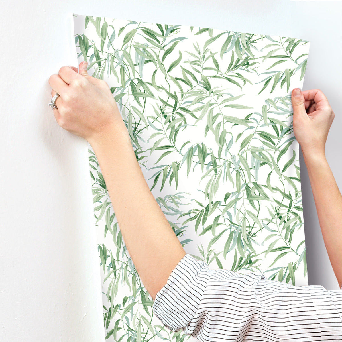 A person with a striped shirt is seen from the shoulders down, carefully aligning a sheet of York Wallcoverings Willow Grove Sky Wallpaper Blue (60 Sq.Ft.) with a green leafy pattern against a white wall. The person's hands and part of their arms are visible, and they are wearing a ring on one finger.