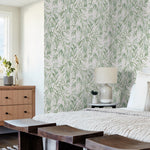 A bedroom with a light and airy design features a bed with a light-colored woven blanket, wooden benches at the foot, and a bedside table with a lamp and potted plant. A dresser, small statue, and vase with flowers sit near a window. The walls are adorned with York Wallcoverings Willow Grove Sky Wallpaper Blue (60 Sq.Ft.) featuring leafy patterns.