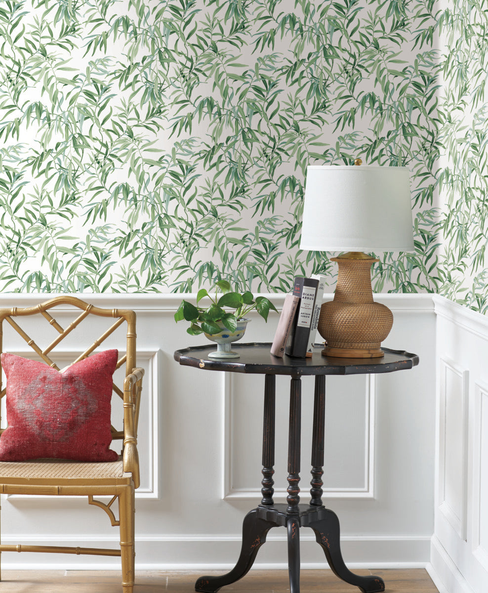 A cozy corner featuring a wooden chair with a red cushion, a dark round side table holding a beige woven lamp and a small potted plant, set against York Wallcoverings' Willow Grove Sand Wallpaper Beige, Grey (60 Sq.Ft.) with white wainscoting underneath, adding natural elegance to the space.