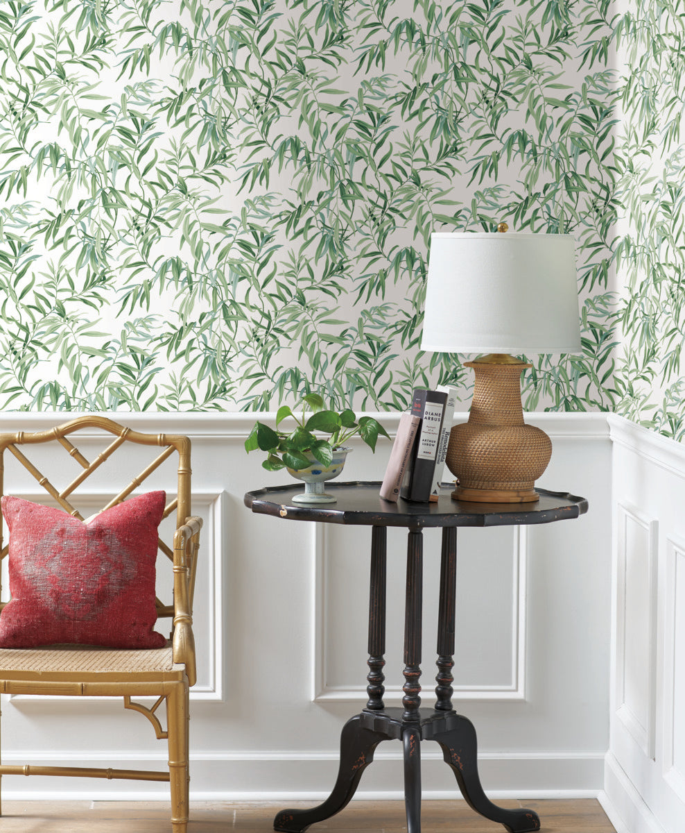 A corner of a room with green leafy **Willow Grove Clay Wallpaper Pink (60 Sq.Ft.) by York Wallcoverings** and white wainscoting. A rattan chair with a red cushion is beside a small black side table. The table holds a wicker lamp with a white shade, a potted plant, and a few books, creating the perfect botanical retreat on light wood floors.