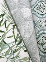 Three rolled-up pieces of wallpaper are displayed. The designs include leafy green vines on white, intricate white leaves on a gray background, and a teal and white ornate pattern with floral and geometric elements, showcasing the natural elegance of Willow Grove Sand Wallpaper Beige, Grey (60 Sq.Ft.) featuring SureStripª substrate by York Wallcoverings.