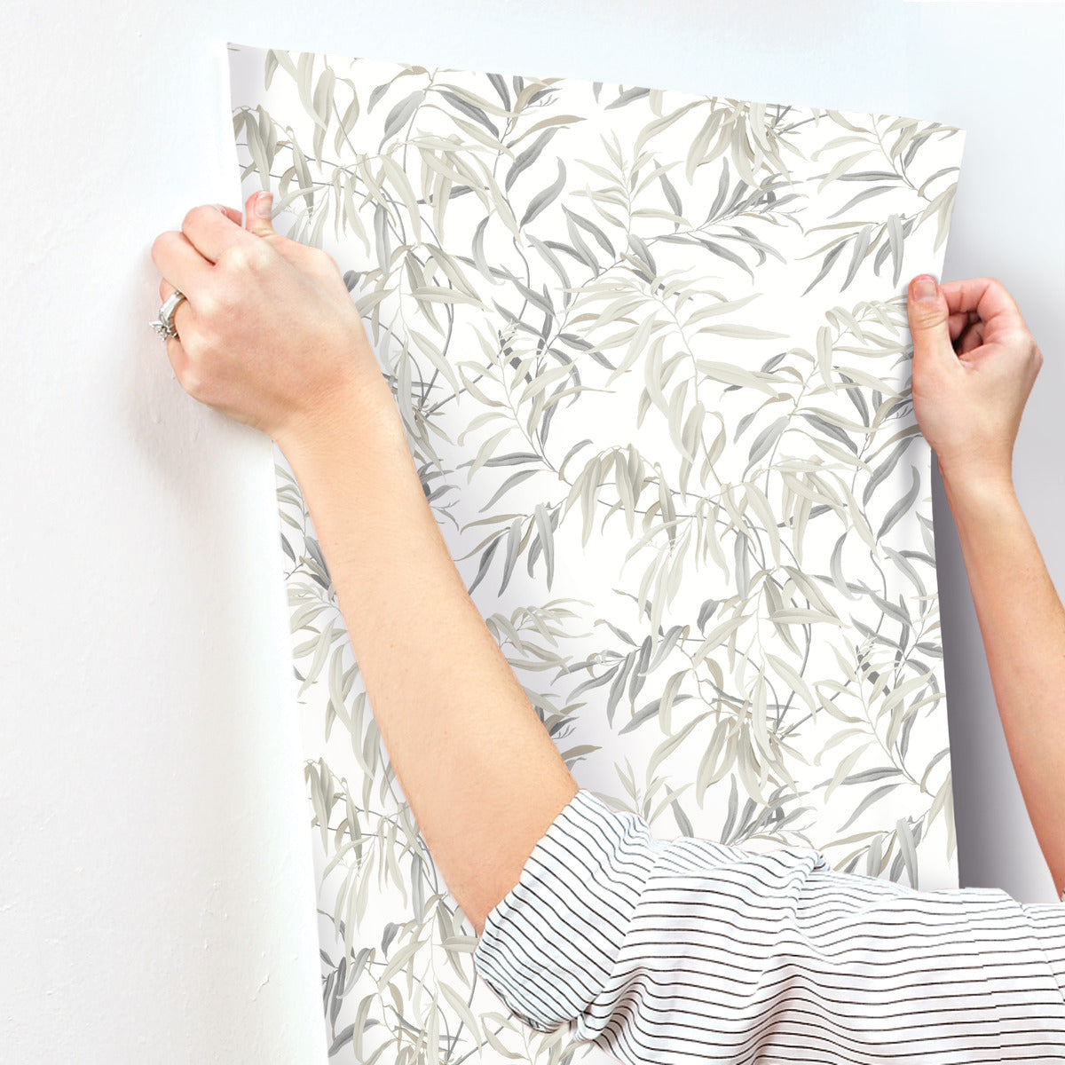 A person wearing a striped shirt is applying York Wallcoverings Willow Grove Clay Wallpaper Beige(60 Sq.Ft.) with a leafy pattern to a white wall. Their hands are smoothing out the high-performance substrate, and a ring is visible on one finger. The wallpaper features light, delicate leaves in gray tones reminiscent of willow trees.