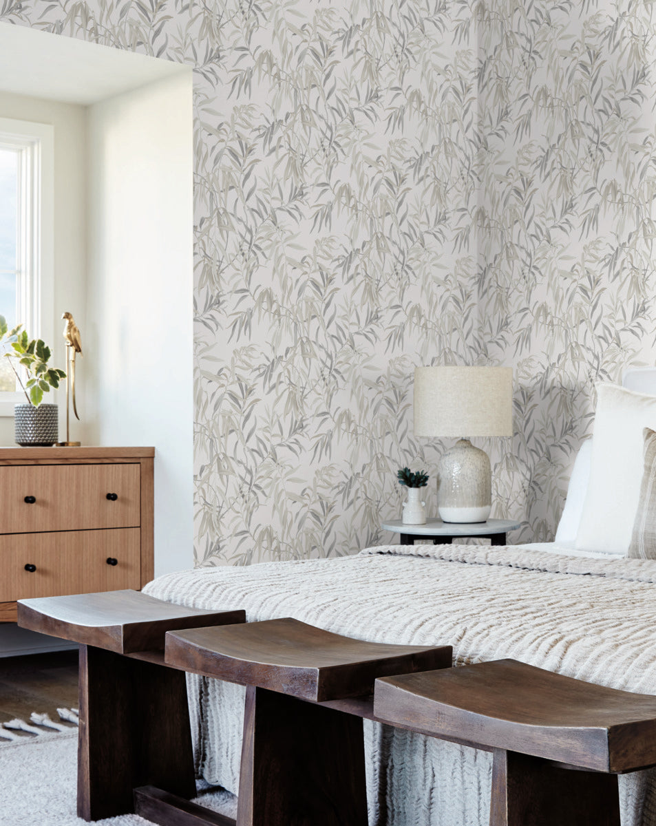 A cozy bedroom with a bed adorned with a knitted blanket and pillows. A bedside table holds a lamp and a small plant. The York Wallcoverings Willow Grove Sand Wallpaper Beige, Grey (60 Sq.Ft.) features a leafy pattern, adding natural elegance to the space, while a wooden dresser sits under the window on the left. Three wooden benches are placed at the foot of the bed.