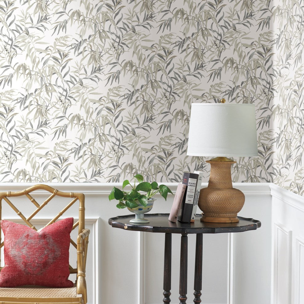 A room with York Wallcoverings Willow Grove Clay Wallpaper Pink (60 Sq.Ft.) and white wainscoting features a wooden chair with a red cushion, a dark wooden table holding a wicker-base lamp with a white shade, a potted green plant, and two books.