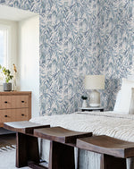 A cozy bedroom features York Wallcoverings Willow Grove Sky Wallpaper Blue (60 Sq.Ft.) with blue and white leafy designs. The room includes a wooden dresser topped with a plant and gold statue, a bed with a cream-colored comforter, a bedside table with a lamp, and wooden stools at the foot of the bed.