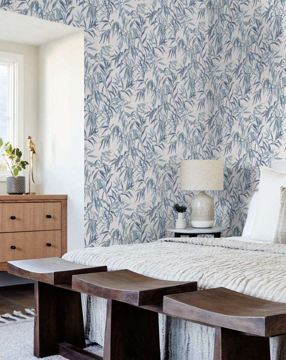 A cozy bedroom features York Wallcoverings Willow Grove Sand Wallpaper Beige, Grey (60 Sq.Ft.) with gray and white foliage, adding a touch of natural elegance. A wooden bench sits at the foot of a neatly made bed, adorned with layered blankets and pillows. The room includes a wooden dresser with a potted plant and a lamp on a bedside table.