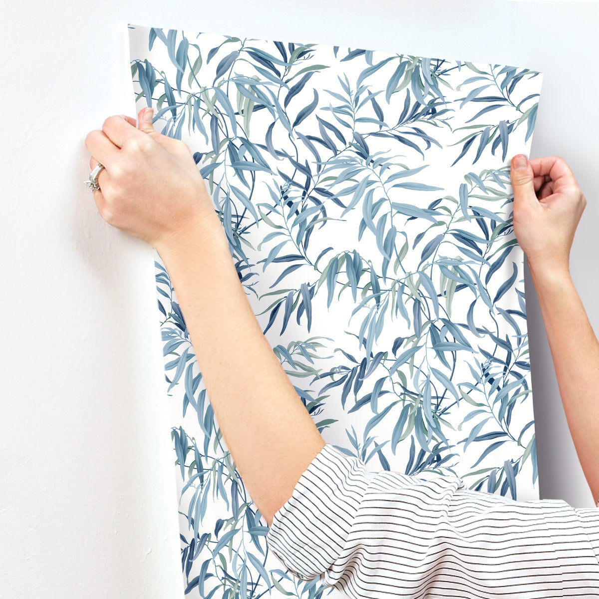 A person is applying a sheet of York Wallcoverings Willow Grove Sky Wallpaper Blue (60 Sq.Ft.) with a leafy, teal, and blue botanical pattern to a white wall. The person, wearing a striped long-sleeve shirt and a ring on their left hand, demonstrates the effortless charm of this easy installation wallpaper.