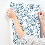 A person with a striped shirt is seen applying York Wallcoverings Willow Grove Sand Wallpaper Beige, Grey (60 Sq.Ft.) with a leafy blue and green plant design to a white wall. Showcasing natural elegance, the person's hands are visible, holding the SureStrip™ substrate in place.