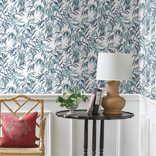 A small round black table holds a wicker lamp with a white shade, a plant, and two books against a wall adorned in York Wallcoverings Willow Grove Sky Wallpaper Blue (60 Sq.Ft.) featuring green leaf patterns. A wooden chair with a red cushion sits next to the table, and white wainscoting adorns the lower part of the wall.