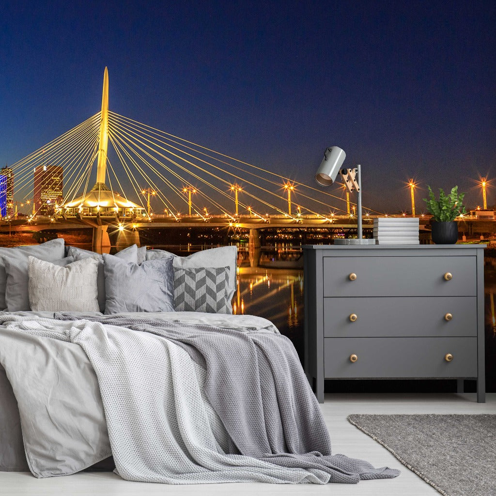 A cozy bedroom setup with a bed covered in grey linens fronting a large window displaying a night landscape of a cityscape with a lit Decor2Go Wallpaper Mural featuring the Winnipeg Bridge.