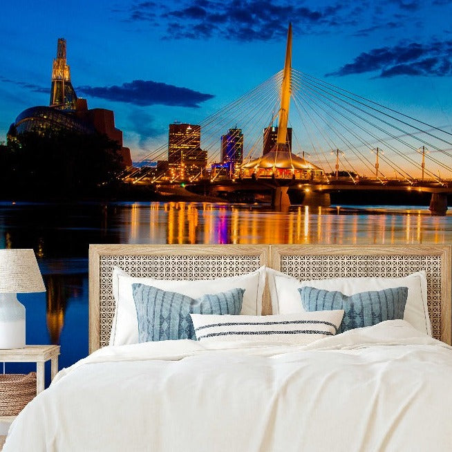 A luxurious bedroom with a bed facing a large window showcasing a scenic view of a "Decor2Go Wallpaper Mural," featuring the striking cable-stayed bridge and illuminated buildings reflecting on a river.