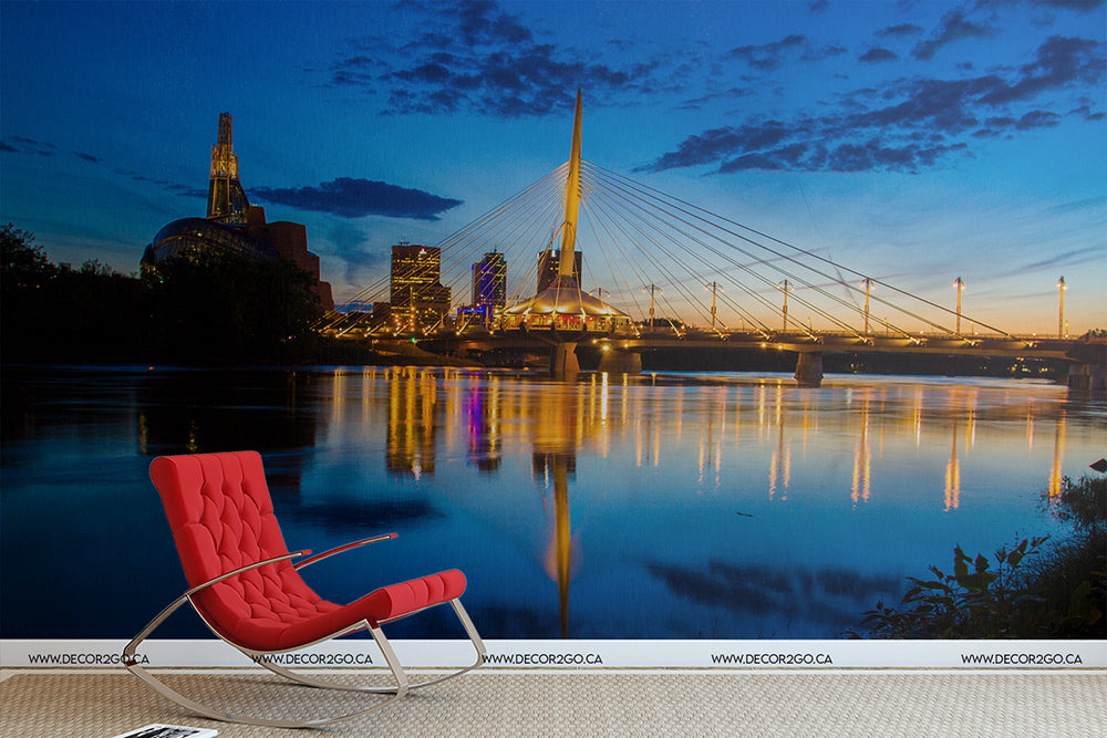 A modern red lounge chair sits by a calm river at twilight, facing a cityscape with a striking cable-stayed bridge lit up against the evening sky. Gently illuminated buildings reflect on the water's Winnipeg Twilight Wallpaper Mural from Decor2Go Wallpaper Mural.