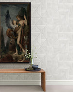 A framed painting depicting a mythological scene hangs on Cheverny Geometric Wood and Gold Wallpaper (56 SqFt) by York Wallcoverings. Below it, a wooden bench displays a transparent vase with delicate white flowers next to a tall white candle in a dark holder. The corner of a beige floor is visible.
