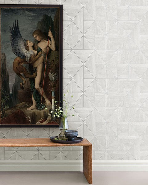 A framed painting depicting a mythological scene hangs on Cheverny Geometric Wood and Gold Wallpaper (56 SqFt) by York Wallcoverings. Below it, a wooden bench displays a transparent vase with delicate white flowers next to a tall white candle in a dark holder. The corner of a beige floor is visible.