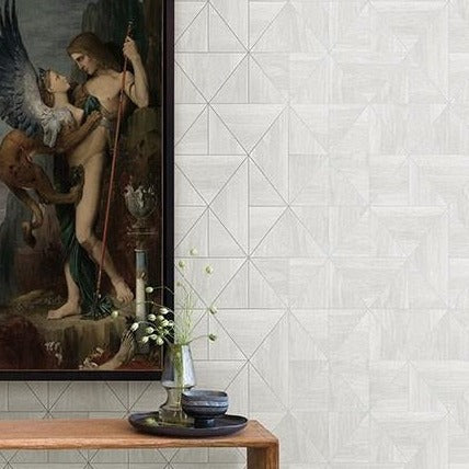 A painting of a mythical scene featuring a winged creature and human is displayed on a wall adorned with York Wallcoverings' White and Silver Cheverny Geometric Wood Wallpaper. Below, aligning with the modern rustic aesthetic, a wooden table supports a vase of white flowers and two small bowls.