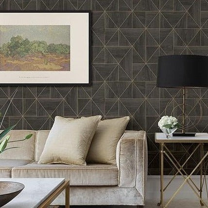 A modern living room features a plush beige sofa with two cushions, a gold-framed side table with a black lamp, and a small white flower arrangement. The wall has Cheverny Geometric Wood and Gold Wallpaper (56 SqFt) by York Wallcoverings and a framed landscape painting above the sofa.